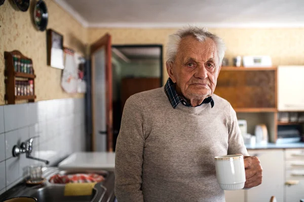 Portret van een oudere man met een kop thee die thuis staat, kijkend naar de camera. — Stockfoto