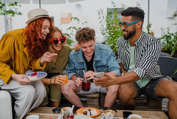 Grupa szczęśliwych młodych ludzi ze smartfonem siedzi w kawiarni na świeżym powietrzu na wycieczkę, rozmowy. — Zdjęcie stockowe