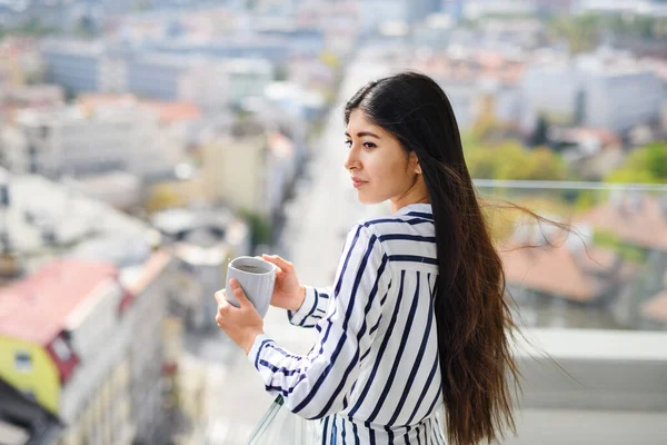 Porträt einer jungen Frau mit Kaffee, die auf einem Balkon in der Stadt steht und das Stadtbild betrachtet. — Stockfoto