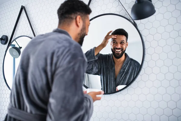 Young man applying cream on face indoors at home, morning or evening routine concept. — Stock Photo, Image