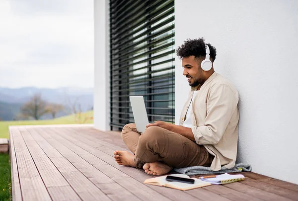 Młody człowiek student z laptopem i słuchawkami siedzi na patio w domu, studiuje. — Zdjęcie stockowe