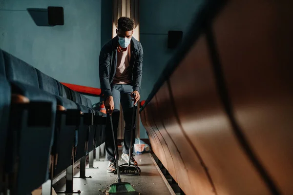 Молодой человек уборщик подметает пол в кинотеатре после фильма, коронавирусная концепция. — стоковое фото