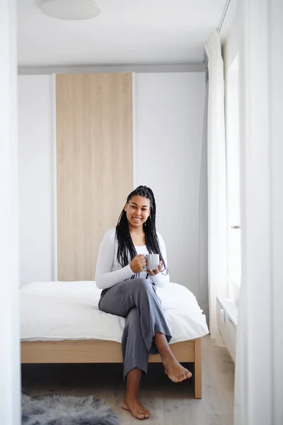 Szczęśliwa nastolatka z filiżanką herbaty siedzi w sypialni w domu, patrząc na aparat. — Zdjęcie stockowe
