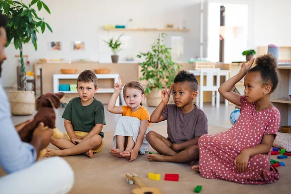 Gruppe kleiner Kindergartenkinder mit einem männlichen Lehrer, der drinnen im Klassenzimmer auf dem Boden sitzt und die Hände hebt. — Stockfoto