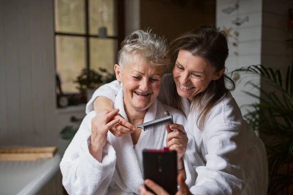 Felice madre anziana in accappatoio con figlia adulta prendendo selfie al chiuso a casa, concetto di auto-cura. — Foto Stock