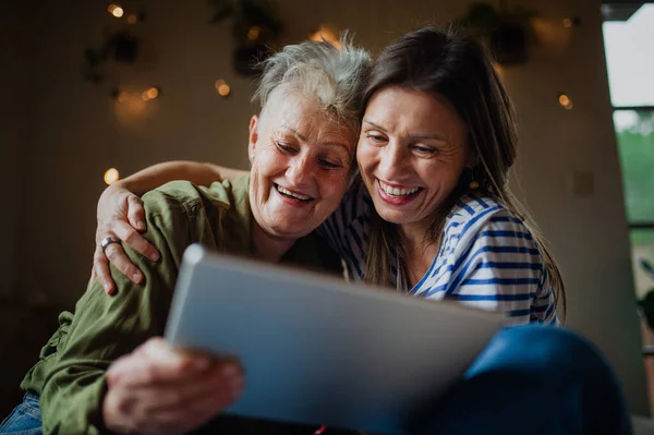 Retrato de mãe sênior feliz com filha adulta dentro de casa, usando tablet. — Fotografia de Stock