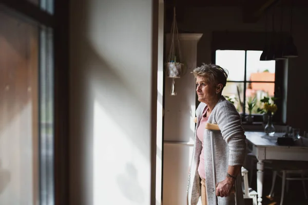 Mujer mayor triste con muletas en el interior de casa, mirando por la ventana. — Foto de Stock