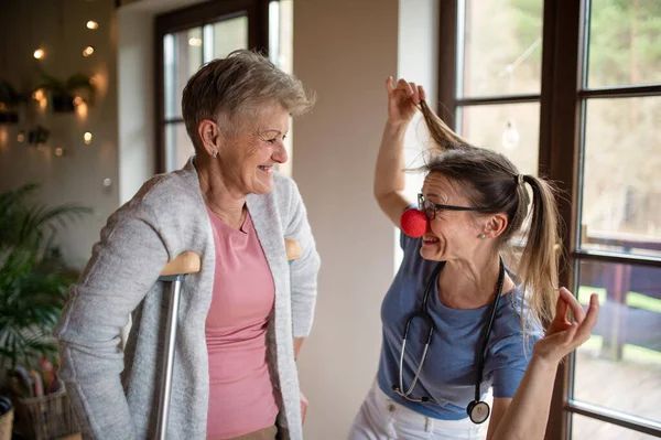 Healthcare worker or caregiver with red nose visiting senior woman indoors at home, having fun. — Stock Photo, Image
