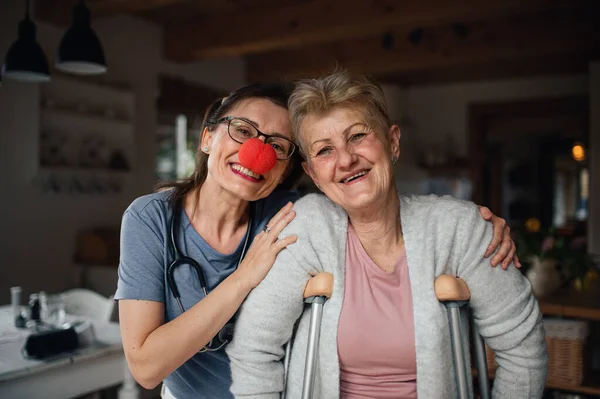 Pracownik służby zdrowia lub opiekun z czerwonym nosem odwiedzający starszą kobietę w domu, patrząc w kamerę. — Zdjęcie stockowe