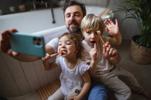 Padre con dos niños pequeños cepillándose los dientes en casa, tomando selfie. — Foto de Stock