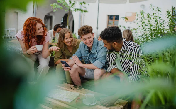 Група щасливих молодих людей зі смартфоном, що сидить у відкритому кафе під час мандрівки містом, розмовляє . — стокове фото