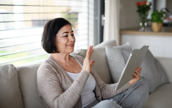 Gelukkig senior vrouw met tablet zitten op de bank binnen thuis, video call concept. — Stockfoto