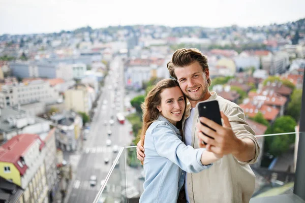 幸福的年轻夫妇在户外的阳台上站着，用智能手机自私自利. — 图库照片
