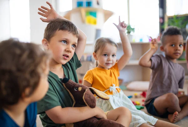 Groep kleine kleuterschoolkinderen die binnen in de klas op de vloer zitten en handen opsteken. — Stockfoto