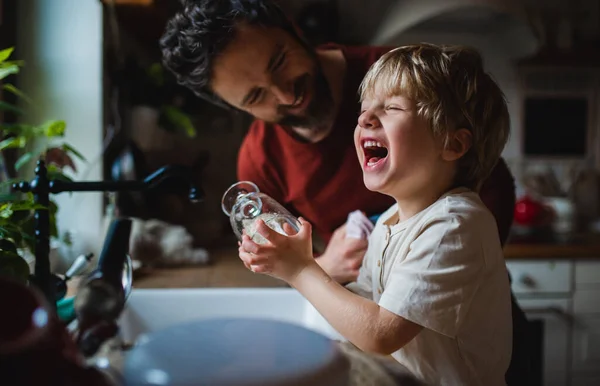 Pequeño niño riendo con el padre divirtiéndose al lavar los platos en el interior de casa, concepto de tareas diarias. — Foto de Stock