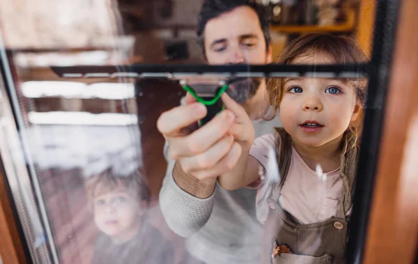 Ragazza felice con padre che pulisce le finestre a casa, concetto di faccende quotidiane. Girato attraverso il vetro. — Foto Stock