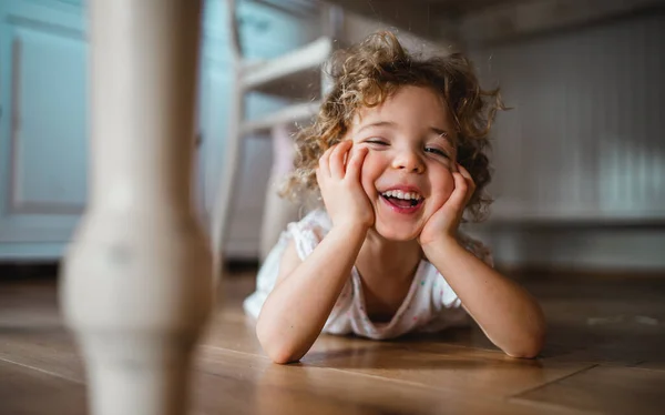 Retrato de linda niña pequeña acostada en el suelo debajo de la mesa en el interior de casa, mirando a la cámara. —  Fotos de Stock