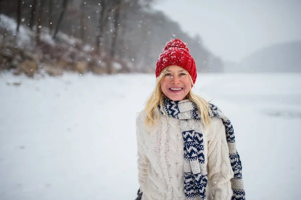 Karlı doğada durmuş kameraya bakarken şapkası açık havada duran mutlu yaşlı kadının ön manzara portresi.. — Stok fotoğraf