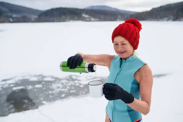 Donna anziana attiva in costume da bagno bere tè all'aperto in inverno, concetto di terapia del freddo. — Foto Stock
