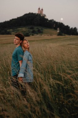 Genç bir çift gün batımında doğa yürüyüşünde kırsalda sırt sırta durup kameraya bakıyor..