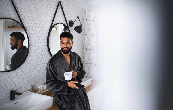 Fiatal férfi kávé és fürdőköpeny beltéri fürdőszobában otthon, okostelefon. — Stock Fotó