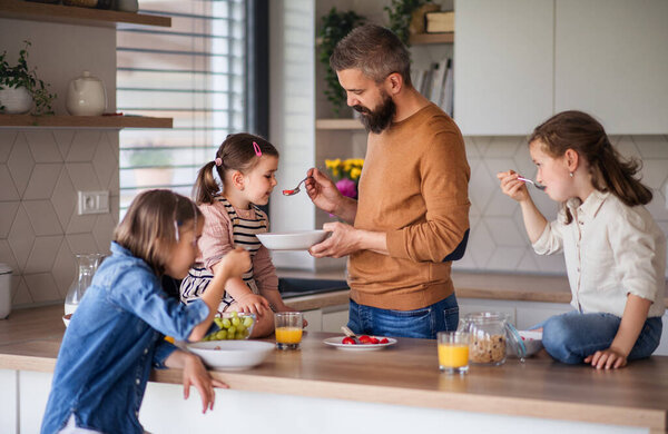 Father with three daughters indoors at home, eating breakfast in kitchen.