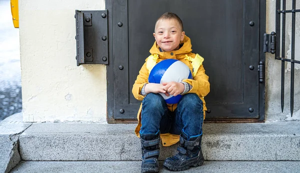 Happy Down menino síndrome com bola ao ar livre sentado na frente da porta, olhando para a câmera. — Fotografia de Stock