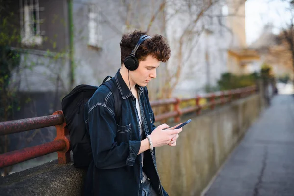 Portret młodego człowieka dojeżdżającego do pracy ze słuchawkami stojącymi na świeżym powietrzu w mieście, za pomocą smartfona. — Zdjęcie stockowe