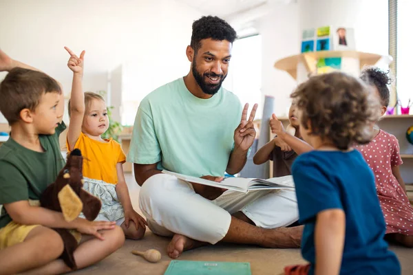 Groep van kleine kleuterschoolkinderen met mannenleraar zittend op de vloer binnen in de klas, met les. — Stockfoto