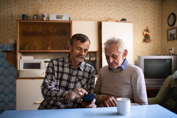 Retrato de homem com pai idoso sentado na mesa dentro de casa, usando smartphone. — Fotografia de Stock