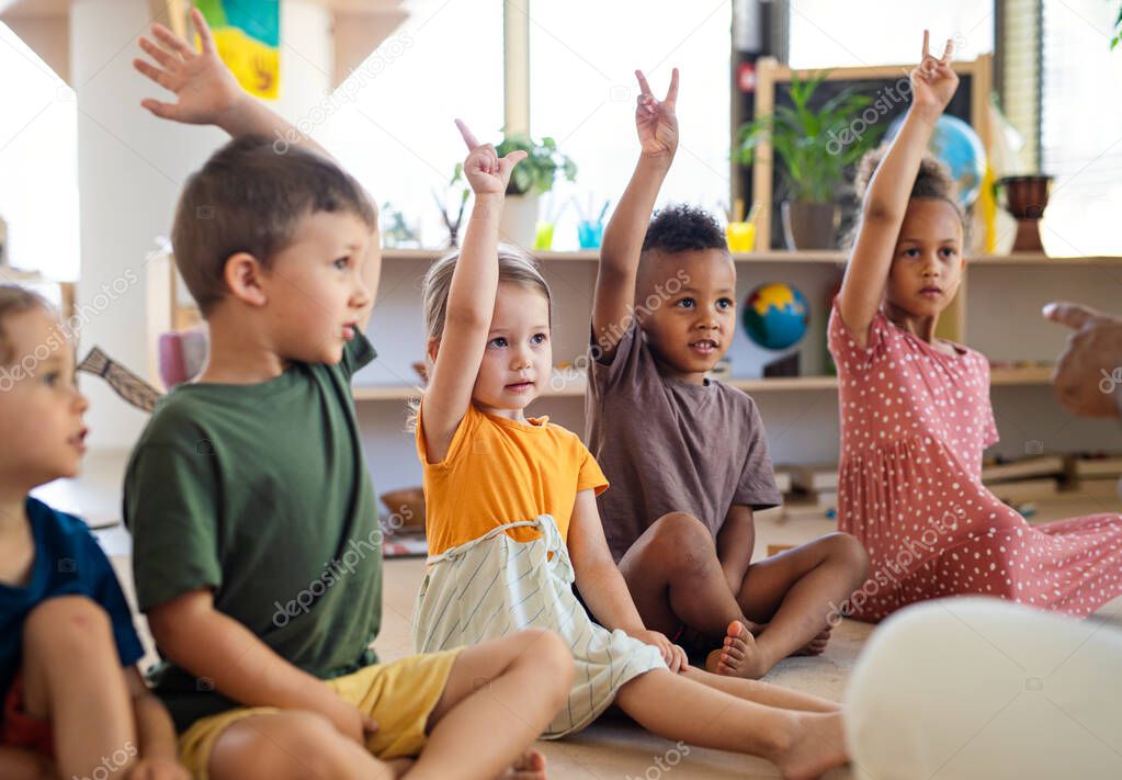 Group of small nursery school children sitting on floor indoors in classroom, raising hands.