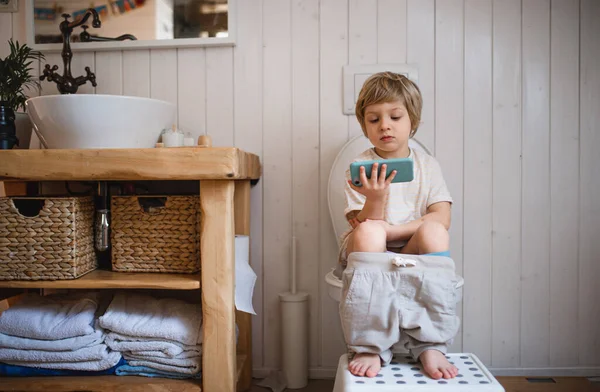 Retrato de lindo niño pequeño sentado en el inodoro en el interior de casa, utilizando el teléfono inteligente. —  Fotos de Stock