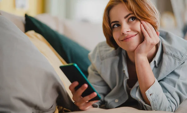 Retrato de mulher jovem usando smartphone, deitado no sofá em casa, olhando para o lado, conceito de redes sociais. — Fotografia de Stock