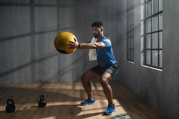 Ung afroamerikansk idrottsman stående och lyfta en medicinboll inomhus, crossfit träningskoncept. — Stockfoto