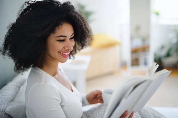 Porträt einer jungen Frau mit Buch und Kaffee drinnen, die es sich im Bett gemütlich macht. — Stockfoto