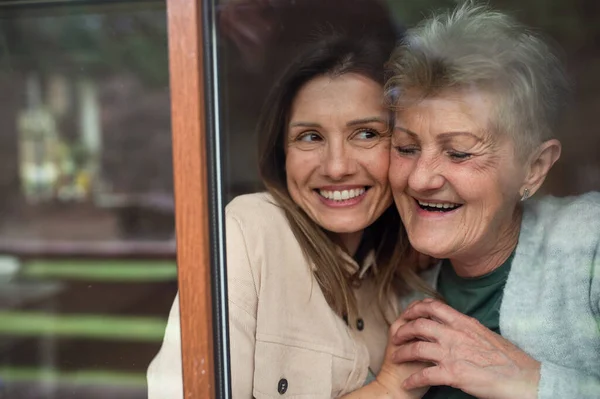 Close-up portrait of happy senior mother with adult daughter indoors at home, hugging. Shot through window. — Stock Photo, Image