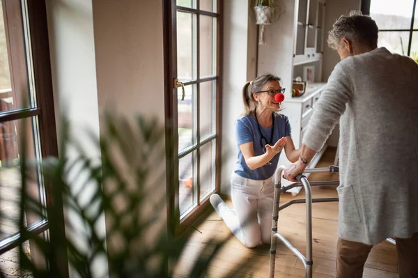 Trabajador de la salud o cuidador con nariz roja visitando a una mujer mayor en casa, divirtiéndose. — Foto de Stock