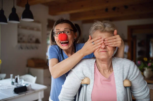 Gezondheidszorgverlener of verzorger met rode neus die bejaarde vrouw thuis bezoekt, plezier heeft en haar ogen bedekt. — Stockfoto