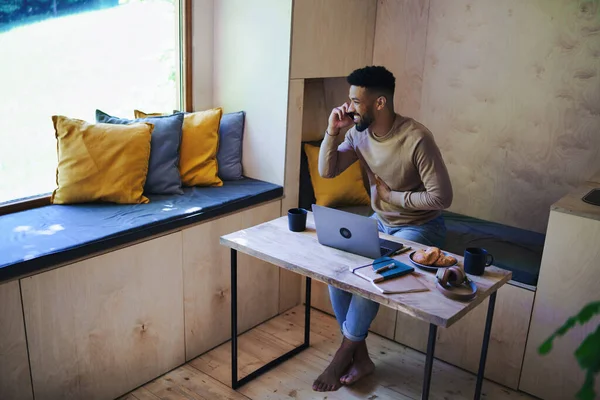 Joyeux jeune homme avec ordinateur portable et smartphone assis, reposant à l'intérieur dans une cabane dans les arbres, week-end et concept de bureau à distance. — Photo