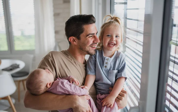 Счастливый молодой человек заботится о своем новорожденном ребенке и маленькой дочери в помещении дома, глядя в камеру. Отцовский отпуск. — стоковое фото