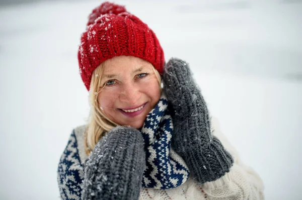 Widok z przodu portret szczęśliwej seniorki z kapeluszem na zewnątrz stojącej w zaśnieżonej naturze, patrzącej w kamerę. — Zdjęcie stockowe