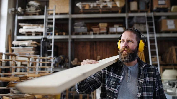 Carpinteiro macho maduro com protetores auditivos carregando placa de madeira, dentro de casa na oficina de carpintaria. Conceito de pequenas empresas. — Fotografia de Stock
