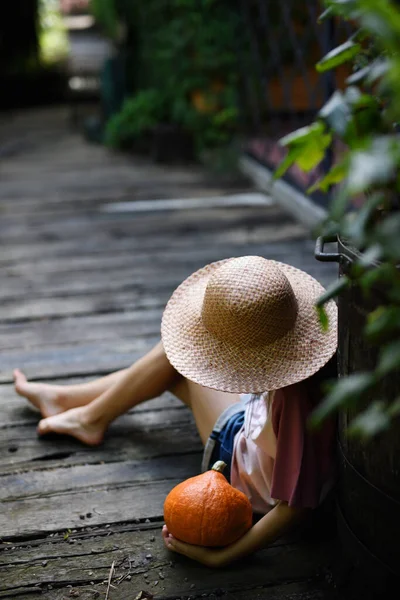 Vista de ángulo alto de niña irreconocible en sombrero sentado y sosteniendo calabaza orgánica al aire libre en la granja. —  Fotos de Stock