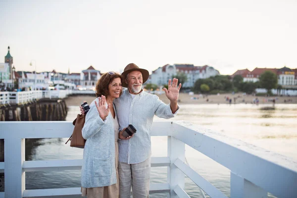 Glada äldre par vawing utomhus på piren vid havet, poserar för foto. — Stockfoto