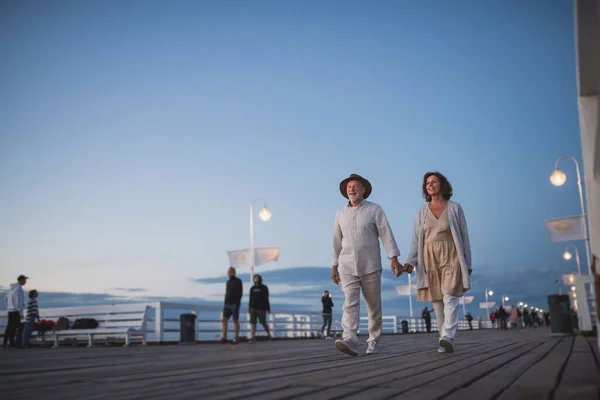 Visão de ângulo baixo de casal sênior feliz andando ao ar livre no cais pelo mar ao entardecer, de mãos dadas. — Fotografia de Stock