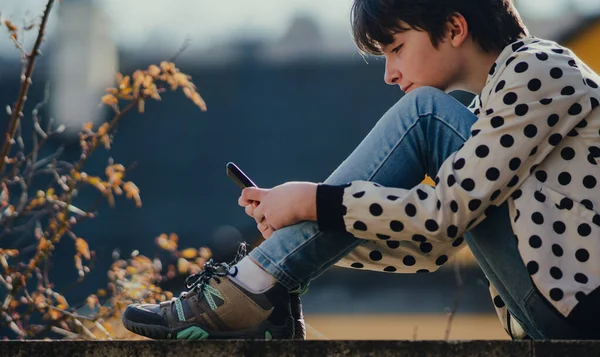 街の中で屋外に座っているスマートフォンを持つ少女のサイドビュー. — ストック写真