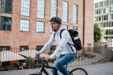 Kasklı ve sırt çantalı bir adam işe bisikletle gidiyor.