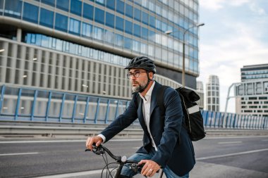 Kasklı ve sırt çantalı bir adam işe bisikletle gidiyor.