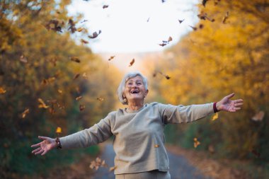 Sonbahar doğasında dışarıda yürüyen yaşlı bir kadın, yere düşen yaprakları havaya fırlatıyor..