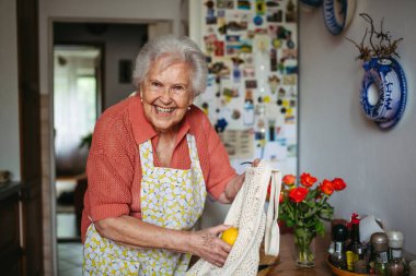 Önlüklü yaşlı bayan evde taze meyve hazırlıyor, elinde de kullanılabilir çanta var..